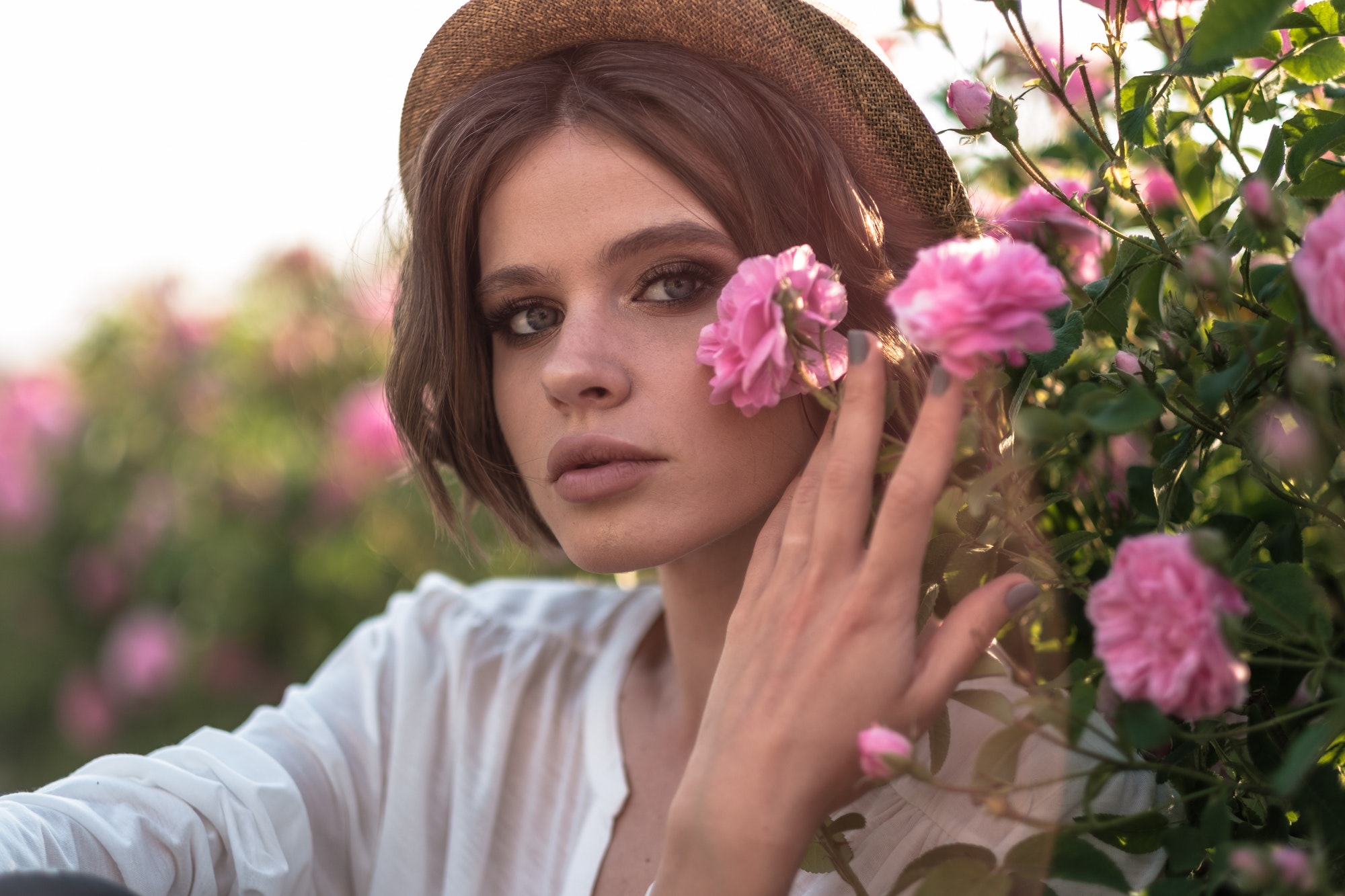 Beautiful woman with curly hair posing near roses in garden. The concept of perfume advertising